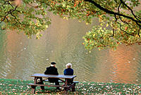 Seniors on a park bench enjoy fall colors reflected in the water, Ontario, Canada  Early autumn, Maple tree overhead will shed all leaves in a week or...