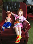 A three year girl with a dolly sits in her grandfather´s chair during a visit.