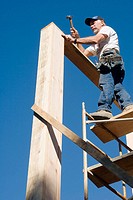 Carpenter standing on scaffolding hammering a nail into a sill plate to connect it to the corner stud of a two-story room.