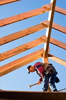 Carpenter hammering trimmer to center post while standing below the framing of the roof rafters at a construction site.