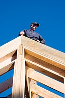 Carpenter standing on the peak of a framed in roof, looking down the rafters at a construction site.