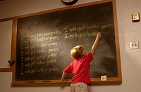 Young blond boy; red t-shirt; at wood-framed classroom blackboard writing ´I will get better grades´multiple times. Light highlighting blackboard and ...