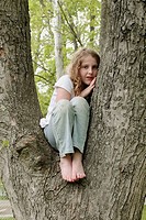 Preteen girl with wavy gold-brown hair and freckles, sitting in tree, in faded jeans and white short-sleeved top, knees close to body and leaning on h...
