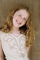 Preteen girl with long gold-brown hair and freckles, smiling at camera, head tilted to side, waist up, against brown wood background