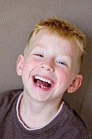 A five-year-old blonde, blue-eyed boy is laughing, showing a gap where he has lost a tooth. He is wearing a brown t-shirt and is standing in front of ...