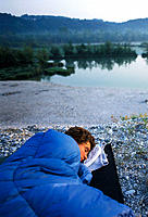 Morning dawn over young woman in sleeping bag at the shore of the reservoir lake of the Isar river near Icking, Upper Bavaria, Germany