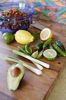 Scallions, lemons, limes, chilis, avocado, on chopping block. Bowl of diced veggies in background.
