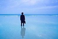 Man standing on the beach in black hat and overcoat with briefcase looks out to the ocean, his image reflected in the wet sand.