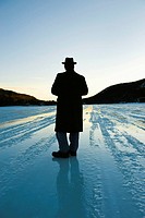 Silhouette of a man wearing an overcoat and a hat standing on a frozen lake strating off to the horizon, his back is to the camera.