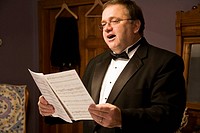 A man wearing a black tuxedo and bow tie singing from a book of sheet music in his dressing room.