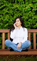 Young Colombian woman with long black hair white shirt and blue jeans, sits cross-legged on a wooden bench while talking on a mobile phone. She is loo...