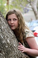 13-year-old girl with blonde-brown long hair and freckles, half leaning against, half hiding behind a tree, up-to-something expression