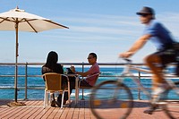 A couple sit at a beachfront table on Las Canteras Beachfront as a policeman cycles past