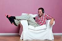 Male in casual clothing relaxing on an armchair wearing headphones whilst watching and listening to a music video on his media player
