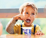 Little boy is eating yogurt, holding a spoon in his hand, sitting at the table