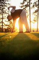 Golfer leaning on driver placing ball on tee with sunlight coming through trees