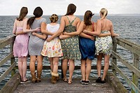 At the end of a pier, six young women with their backs to the camera are connected by arms as they look out into the water