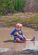 This cute 18 month old Caucasian toddler girl is sitting in her diaper and life vest in a mud puddle and playing as she´s taking off her sandal Great ...