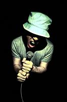 Adult wearing a hat and gray tee shirt male sings in to microphone. Isolated on black background