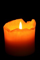 A burning, well used candle with ragged melting edges against a dark background  Isolated