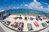 Tourists sunning on beach chairs on the waterfront in Georgetown on Grand Cayman in the Cayman Islands with cruise ships in the caribbean sea off shor...