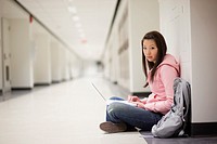 Portrait of a young Asian woman using her laptop computer while sitting on floor in school hallway.