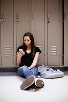 Young Asian woman texting on her smart phone while sitting on floor in school hallway.