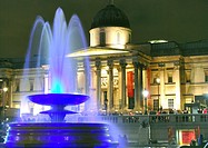 Fountain in Trafalgar Square, London, England, lit up colourfully at night, with the National Gallery illuminated in the background