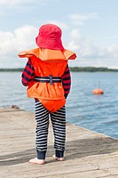 Young child wearing orange life jacket and red hat watching the sea from a wooden jetty