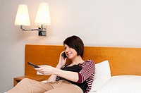 Young woman teenager sitting on bed watching TV and talking on mobile phone