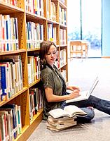 An 18 year old girl sitting on the floor in a library with a laptop computer and books stacked next to her.