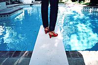 A woman wearing bright red high heel shoes on a diving board in front of a swimming pool.