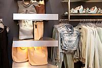 Blouses, shirts and jackets hanging on rack in retail shop interior. Bags, handbags, shoulder bags on shelf.