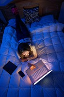 Teenage girl lying on a bed at night working on a laptop computer.