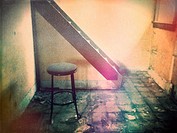 A stool sits on a grungy floor in front of a staircase in a an abandoned house. Ontario, Canada.