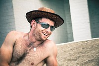 Happy shirtless man in hat and sunglasses relaxing outdoors.