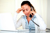 Stressed young woman with headache speaking on phone in front of laptop at workplace.