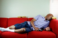 A senior citizen sleeping on a couch with a remote control in hand and potatoes on the couch.