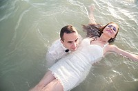 Young man dressed up in a party suit, holding afloat in shalow sea a young woman in a white party dress.