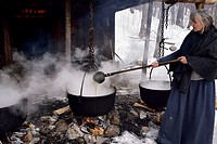 Woman in pioneer clothes ladling evaporated sap in cast iron pots to produce maple syrup.