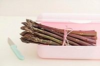 Bunch of fresh purple asparagus in pink plastic container with kitchen knife.
