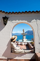 Woman with straw hat relax in an arch with a water fountain over alpine lake Maggiore with snow-capped mountain in a sunny day in Ticino, Switzerland.