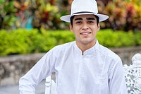 Young hispanic man in formal clothes, wearing white shirt, pants and hat, smiling at camera.