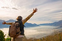 Woman with outstretched arms and backpack enjoy panoramic view with sea of fog over alpine lake Maggiore with mountain in Ticino, Switzerland.