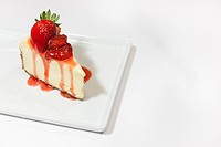 A white cheesecake with dripping strawberry sauce and a graham cracker crust, garnished with whole strawberries on a square plate. Styled with a on a ...