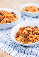 Three bowls of Fusilli pasta, with a tomato meat suace, in white bowls on wodden cutting boards and on a blue checkered napkin.