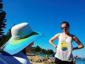 Teenage daughter in a tee-shirt looking at her mother who is wearing a blue and white straw hat while on vacation on a beach on Georgian Bay, Lion´s H...