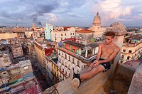 A man sits on the edge of a rooftop with El Capitolio visible in the distance in Central Havana, Cuba.