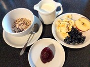 Cereals with oatflakes, fresh blueberries, jam, banana slices half of an apple and a jug with milk