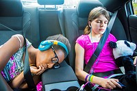 2 girls, one caucasian preteen and one african american teen sucking her fingers in the car sleeping and resting with stuff animals and earphones on.